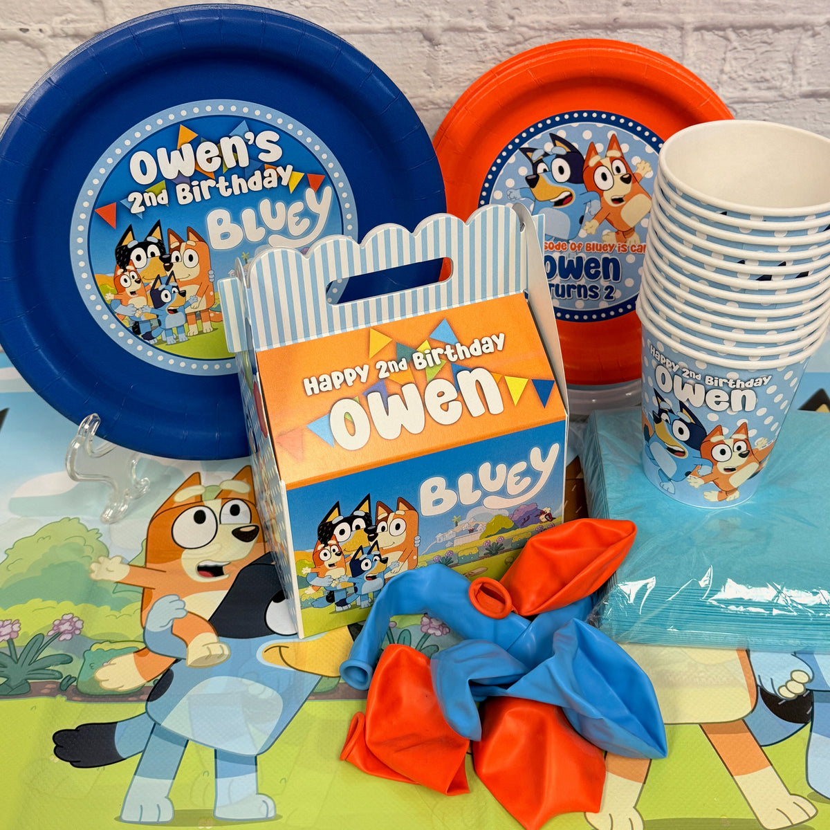 Blue and orange Bluey-themed party supplies including paper plates, cups, a gift box, and balloons personalized for your child's Bluey birthday party.
