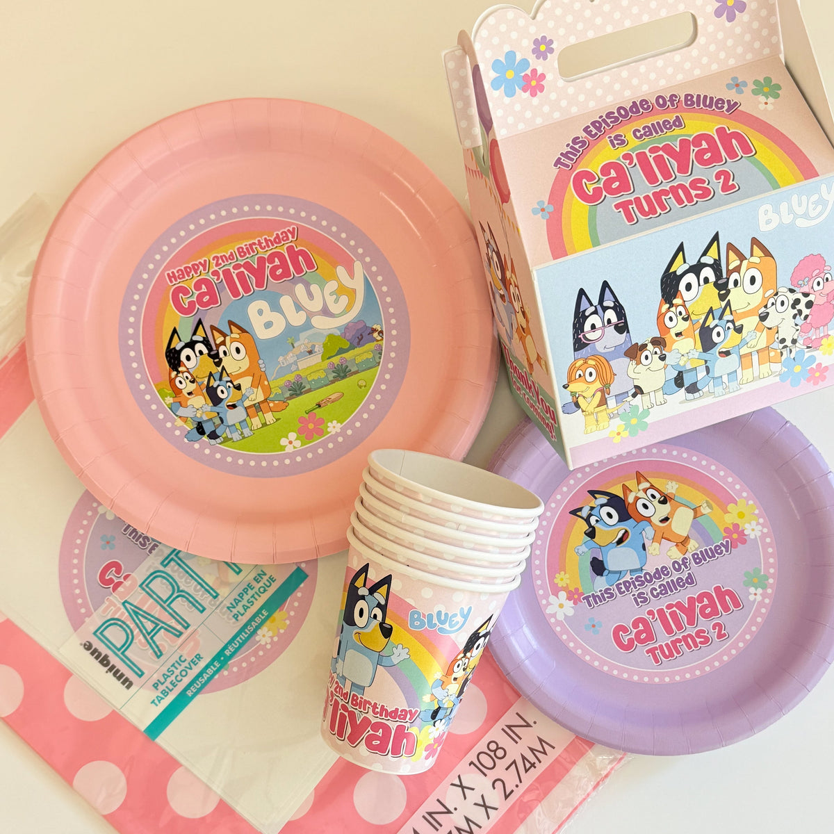 Blue and orange Bluey-themed party supplies including paper plates, cups, a gift box, and balloons personalized for your child's Bluey birthday party. Bluey Girl party, pink with flowers. Pastel Rainbow Bluey party supplies.