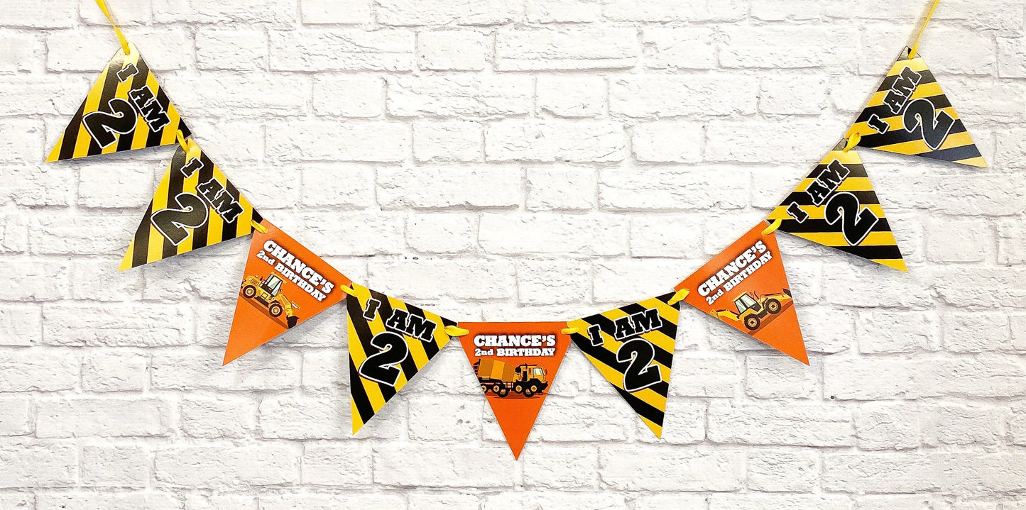 Construction-themed 21st birthday party banner with triangular pennant flags hanging against a white brick wall. The flags feature yellow and black hazard stripes on some and orange on others,  and construction vehicle graphics.
