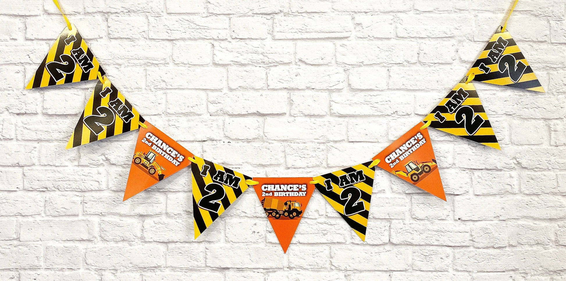 Construction-themed 21st birthday party banner with triangular pennant flags hanging against a white brick wall. The flags feature yellow and black hazard stripes on some and orange on others,  and construction vehicle graphics.